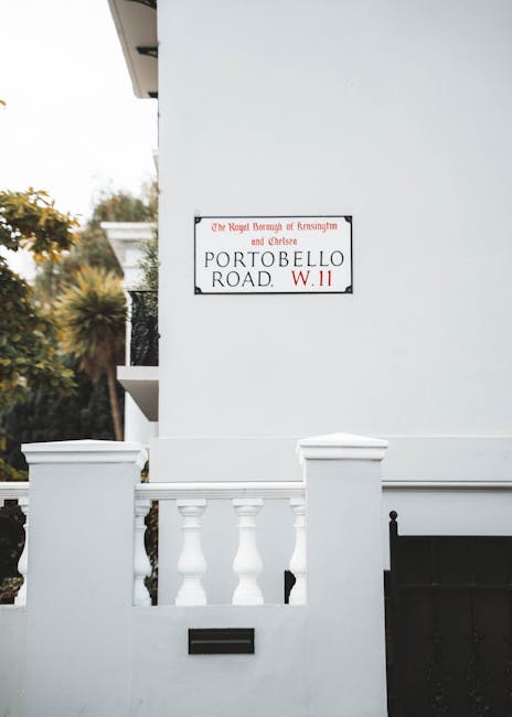 Exterior view of a white residential building showing a section of the front entrance with a low wall and decorative balustrades made of white-painted concrete or stone. Attached to the building's plain white wall is a rectangular street sign reading 'The Royal Borough of Kensington and Chelsea, Portobello Road, W.11' in black and red text. Part of a black metal gate is visible beneath the wall, likely providing access to the property. Surrounding the building, there are trees with green and brown foliage indicating an outdoor setting. The lighting suggests daytime with natural light illuminating the scene. This image is relevant to house removals and moving services, illustrating a typical UK residential street exterior with potential access points for furniture transport and packing activities involved in professional home relocations provided by Man and Van Borough.