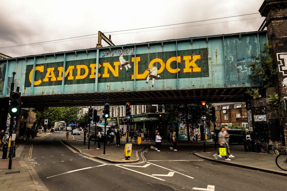 A street scene in Borough featuring a vintage railway bridge with a large, weathered sign reading 'CAMDEN LOCK' in bright yellow and orange letters. The bridge is painted in a faded teal color with visible rust and graffiti. Hanging beneath the bridge are two painted figures, a man and a woman, possibly street art. Below, pedestrians are crossing at a busy intersection with multiple traffic lights, some displaying green signals, and various vehicles, including cars and delivery vans, are visible on the road. On the pavement, there are people walking and cycling, and some are carrying shopping bags. Nearby, there are small street signage and traffic cones, indicating an active urban environment. The scene is well-lit during daytime with an overcast sky, and the surrounding buildings are typical of a London borough area, contributing to an atmosphere of active city life. This setting could relate to home relocations or furniture transport processes managed by companies like Man and Van Borough, often involved in moving and packing logistics.