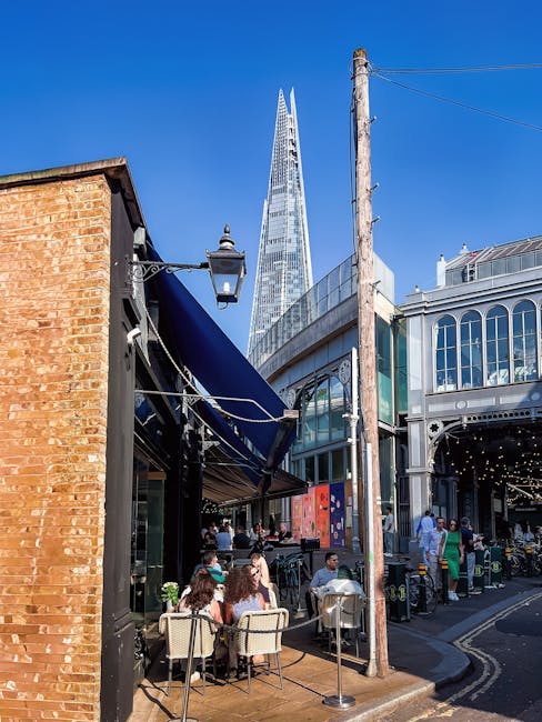 A street scene in Borough Market SE1 during daytime, featuring an outdoor seating area with several tables and chairs occupied by people enjoying food and drinks under a dark blue awning attached to a brick building. Nearby, a tall wooden utility pole with electrical wires stands on the pavement. Adjacent to the seating area is an ornate, glass-fronted building with decorative white framing, housing shops or cafes. In the background, the Shard skyscraper rises prominently against a clear blue sky, emphasizing the urban setting. The overall environment depicts a lively marketplace with foot traffic, and the setup suggests a recent or ongoing home or furniture relocation process involving local removals services, such as those offered by Man and Van Borough, operating in the London area.