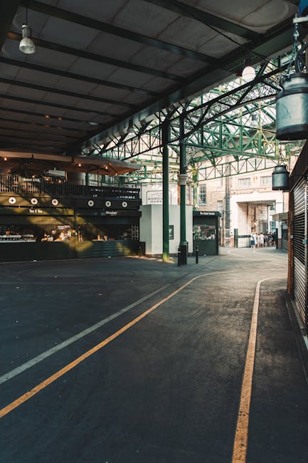 A partially covered outdoor loading area adjacent to a building, with a sloped asphalt surface marked by yellow lines indicating designated parking or loading zones. The space features industrial-style metal structures supporting the roof, which is made of corrugated metal sheets. In the background, there are three visible store signages, including one for a grocery or convenience shop, as well as pedestrians walking along an open sidewalk beyond the loading zone. On the right side, a vehicle, likely a moving van operated by Man and Van Borough, is parked close to the curb, with the rear doors possibly open for loading or unloading furniture, packed boxes, and other household items wrapped in fabric or plastic. The environment is well-lit with natural daylight filtering through the open sides, suggesting a safe and organized setting for home relocation activities, including furniture transport and packing processes associated with house removals in Borough Market SE1.