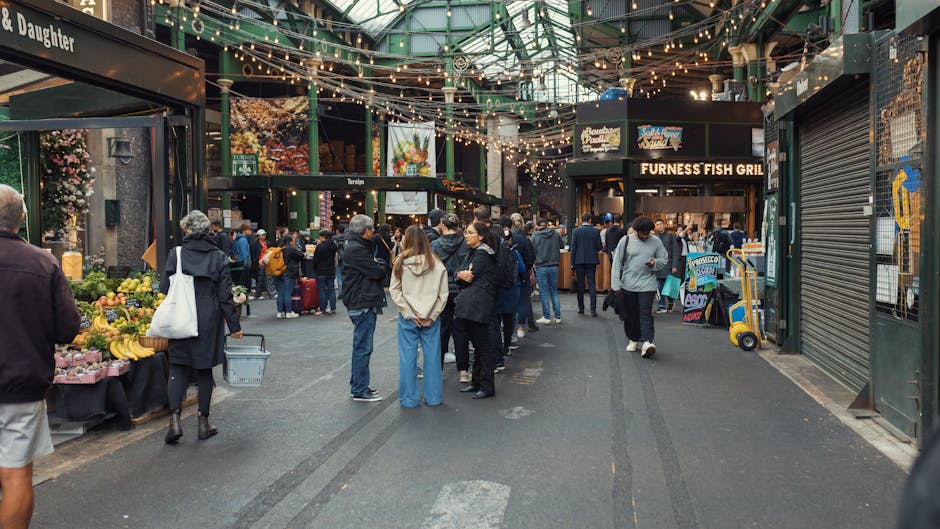 A street scene in Borough Market SE1 during daytime, featuring an outdoor seating area with several tables and chairs occupied by people enjoying food and drinks under a dark blue awning attached to a brick building. Nearby, a tall wooden utility pole with electrical wires stands on the pavement. Adjacent to the seating area is an ornate, glass-fronted building with decorative white framing, housing shops or cafes. In the background, the Shard skyscraper rises prominently against a clear blue sky, emphasizing the urban setting. The overall environment depicts a lively marketplace with foot traffic, and the setup suggests a recent or ongoing home or furniture relocation process involving local removals services, such as those offered by Man and Van Borough, operating in the London area.