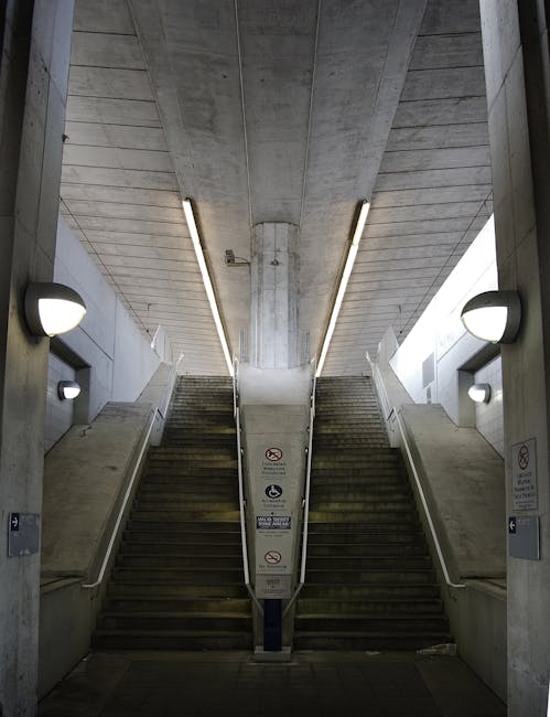 Interior view of a concrete stairwell at London Bridge station with narrow, steep stairs ascending on either side, fitted with white metal handrails along the edges. The stairs are made of dark material, possibly wood or metal, and are positioned between concrete walls with a smooth, grey finish. On the left and right sides, wall-mounted round light fixtures provide illumination. A central metal column supports the ceiling, which features exposed concrete beams with integrated linear lighting. Visible signage and safety notices are affixed to the central column, indicating restrictions, likely related to safety or access. This setting reflects an underground or station environment, suitable for a home relocation or furniture transport process through narrow stair access in Borough, with some equipment or furniture possibly visible nearby as part of a moving service coordinated by Man and Van Borough, specializing in removals and logistics.