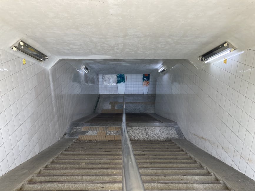 The image shows a downward view of a narrow staircase with concrete steps and a metal handrail in the middle, leading through a tiled underground area possibly part of London Bridge station. The walls are covered with white tiles, some of which are stained or discolored, and the stairwell is lit by four fluorescent light fixtures mounted on the ceiling, two on each side. At the bottom of the stairs, there are partially visible posters on white tiled walls, with an area that appears to be an underpass or corridor. In the foreground, the steps are made of textured concrete, and the staircase looks steep and confined. This setting is indicative of an urban underground transit environment, and the context relates to house or office relocations involving interior stair navigation, as handled by Man and Van Borough, especially in situations with narrow stair access typical of home or building moves.