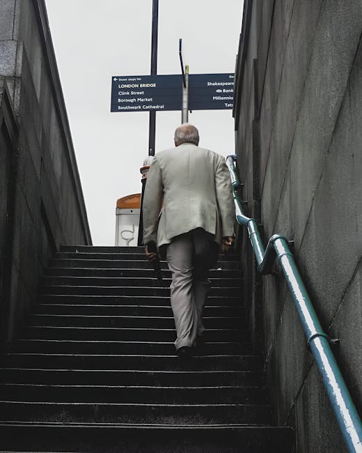 Interior view of a concrete stairwell at London Bridge station with narrow, steep stairs ascending on either side, fitted with white metal handrails along the edges. The stairs are made of dark material, possibly wood or metal, and are positioned between concrete walls with a smooth, grey finish. On the left and right sides, wall-mounted round light fixtures provide illumination. A central metal column supports the ceiling, which features exposed concrete beams with integrated linear lighting. Visible signage and safety notices are affixed to the central column, indicating restrictions, likely related to safety or access. This setting reflects an underground or station environment, suitable for a home relocation or furniture transport process through narrow stair access in Borough, with some equipment or furniture possibly visible nearby as part of a moving service coordinated by Man and Van Borough, specializing in removals and logistics.