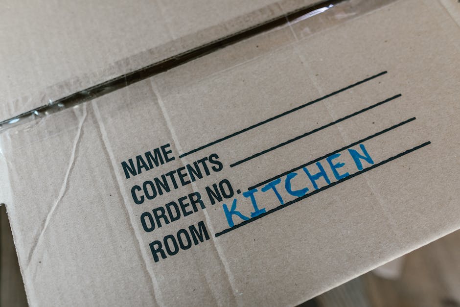 A person preparing for a home relocation within a residential property, partially visible with their torso and arms shown, is carefully lifting a medium-sized cardboard box from a stack of similar box