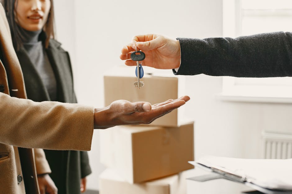 A person in a dark blazer is handing over a set of house keys on a small tray to another individual dressed in a beige coat during a home relocation process inside a well-lit room with white walls and a window. In the background, there are several cardboard boxes and packaging materials stacked on a surface, indicating packing and moving activities. The scene captures the formal handover of keys as part of a house or flat move, with the focus on the keys being transferred, highlighting elements of furniture transport, packing, and the logistical steps involved in house removals, as performed by companies like Man and Van Borough.