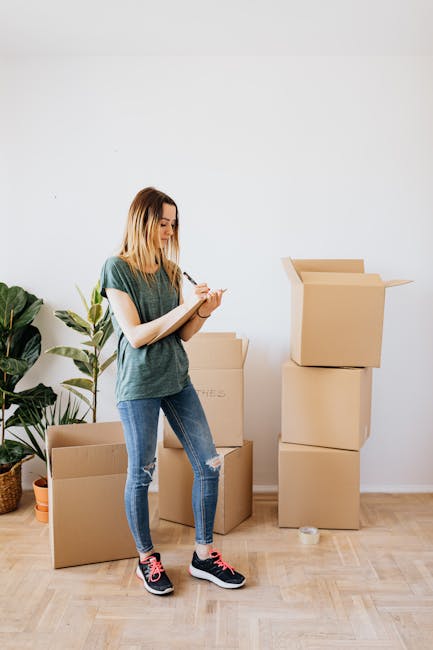A young woman with shoulder-length blonde hair, wearing a casual green T-shirt, ripped blue jeans, and black and pink sneakers, standing indoors on a wooden floor next to several stacked cardboard moving boxes. She is holding a pen and a small notepad, appearing to take notes or check inventory. To her left, there is a large potted plant with broad green leaves, and the background features a plain white wall with no additional decorations. The scene suggests she is preparing for or organizing a home relocation or move, facilitated by professional services such as those offered by Man and Van Borough. The environment indicates a moving process in progress, with a focus on packing and moving logistics.