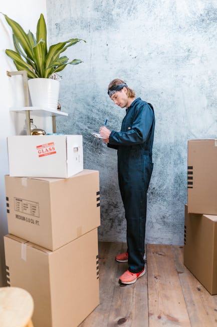 A person in a dark blazer is handing over a set of house keys on a small tray to another individual dressed in a beige coat during a home relocation process inside a well-lit room with white walls and a window. In the background, there are several cardboard boxes and packaging materials stacked on a surface, indicating packing and moving activities. The scene captures the formal handover of keys as part of a house or flat move, with the focus on the keys being transferred, highlighting elements of furniture transport, packing, and the logistical steps involved in house removals, as performed by companies like Man and Van Borough.
