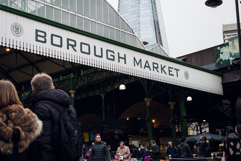 A busy scene outside Borough Market showing multiple pedestrians walking across the pavement and entering the market entrance, which is an ornate, green metal structure with decorative detailing and a