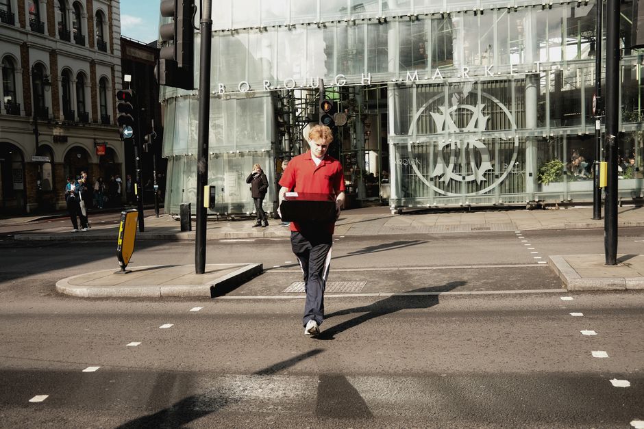 A delivery worker from Man and Van Borough, dressed in a red shirt and dark trousers, is walking across a pedestrian crossing on a city street, carrying a large black box with both hands. Behind him, there is a modern glass building with a circular logo and the words 'BOROUGH MARKET' visible at the top. The street scene includes some pedestrians standing on the sidewalk near the building, traffic lights, and black bollards preventing vehicle entry. The pavement is partially shaded, with sunlight illuminating the worker and surrounding area, suggesting daytime. The scene depicts a moment in the logistics process of packing and transporting office equipment or furniture as part of a house or office removal service.