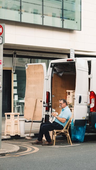 A man in casual clothing, wearing glasses, is seated on a wooden chair on the pavement beside an open white cargo van used for house and office removals. The van's loading area is visible with a large wooden panel leaning against its side, and the cargo space contains cardboard boxes and packing materials ready for transport. The scene is set outside a modern building with large glass windows and a white facade, indicating an urban environment suitable for furniture transport and packing during a home or office relocation. The man appears to be taking a break during the loading process, which involves careful handling of furniture and boxes, supported by moving equipment such as trolleys or straps possibly used elsewhere nearby. Man and Van Borough specializes in removals, including logistics for minimal downtime during office or house moves, as evidenced by the organized loading activity captured in this image.