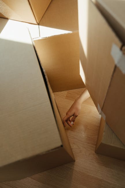 A dimly lit interior room during a house move, with cardboard boxes of various sizes stacked in front of a black upright piano. Some boxes are sealed with packing tape, others are open or partially un