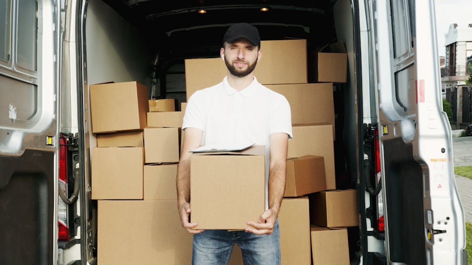 A man in a white polo shirt, dark jeans, and a black cap stands inside the back of a large white moving van, holding a medium-sized cardboard box. The van's interior is filled with numerous stacked cardboard boxes, some wrapped in plastic or fabric for protection during packing and transport. The boxes vary in size and are arranged neatly within the van, which is parked outdoors on a paved area with a residential street visible in the background. The environment suggests a home relocation, with the man preparing to load or unload household items as part of a professional removals service. Man and Van Borough is a company specializing in house removals and furniture transport, supporting seamless packing and moving logistics.