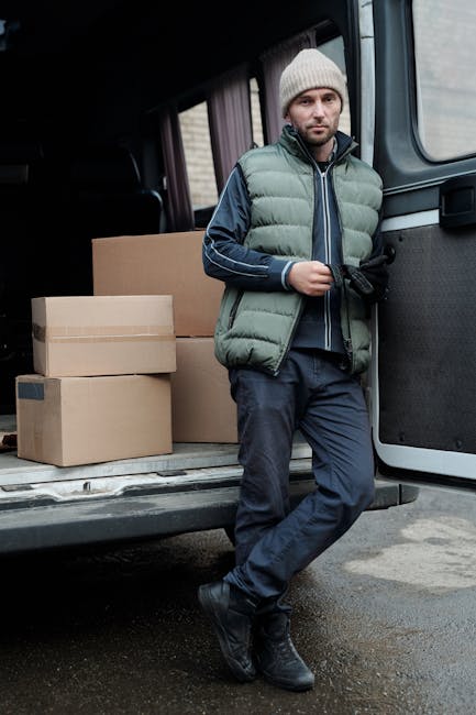 Two movers from Man and Van Borough are engaged in a home relocation process outside a residential property, loading cardboard boxes into the back of a white van. The van is parked on a brick-paved driveway adjacent to a brick wall and a garden area with greenery. One mover, wearing a navy jacket, black quilted vest, and white shoes, is lifting a large cardboard box, while the other, dressed in a dark hoodie, black pants, and sneakers, assists by holding the box. To the left, a black hand truck with two wheels is positioned upright on the pavement, ready to transport additional packing materials. The open rear doors of the van reveal more packed boxes inside, indicating an ongoing packing and moving operation as part of a furniture transport or removal service, highlighting the load preparation and vehicle loading process typical of house removals services.