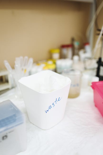 A white paper waste bin labeled 'waste' in blue handwritten text is positioned on a work surface, with various containers and bottles of different shapes and sizes, some with yellow caps and others with white lids, visible in the background. The setting appears to be an indoor workspace or storage area designated for packing or disposal tasks, with a shelf above holding additional containers, possibly for packing materials or cleaning supplies. The surface on which the waste bin sits is covered with a white cloth or paper. This scene reflects the organisation and preparation involved in home relocation and furniture transport, with careful sorting and disposal of packing waste as part of the moving process handled by Man and Van Borough, a professional removals service specializing in house removals and moving logistics.