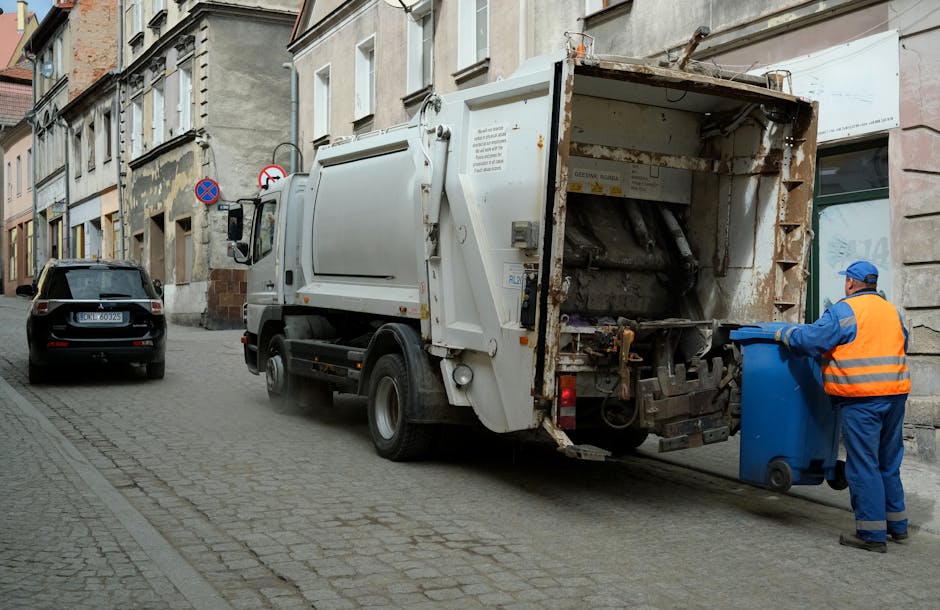 A street scene showing a white garbage collection truck from Man and Van Borough actively engaged in waste disposal, parked parallel to a row of older, multi-story residential buildings with weathered facades. The truck's rear hopper is open, revealing empty compartments for waste collection. A worker dressed in a blue uniform and an orange high-visibility vest is standing beside the truck, completing the loading process by handling a blue wheeled bin, which is positioned on the cobblestone pavement adjacent to the truck. The worker is in the act of tipping or retrieving the bin, contributing to the home relocation and packing process associated with removals and rubbish clearance. Next to the worker, another vehicle, a grey SUV, is parked facing away from the truck, and the street is narrow with minimal traffic, consistent with urban residential areas. The scene is lit by natural daylight, emphasizing the equipment, materials such as plastic bins, and the logistics involved in household waste removal, aligning with Southwark Council's bulky waste rules for removals and disposal, as managed by Man and Van Borough.