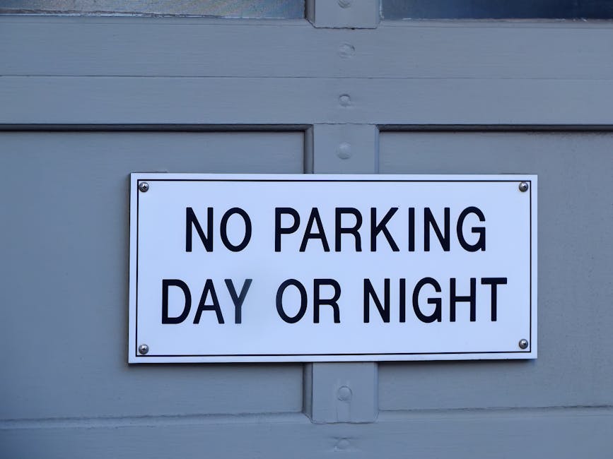 A rectangular white metal sign with black uppercase text stating 'NO PARKING DAY OR NIGHT' is mounted on a light grey wooden garage door. The sign is secured with four screws, one at each corner. The garage door features horizontal panels separated by narrow, slightly darker grooves, and is painted in a matte finish. Natural light illuminates the scene, revealing the texture of the wood and the clean, sharply printed lettering on the sign. This setting relates to house removals and parking regulations as part of the logistical considerations involved in home relocation and furniture transport, with [COMPANY_NAME] such as Man and Van Borough often navigating these regulations during the loading process within the circumstances outlined by Southwark Council rules for Borough removals and parking permits.