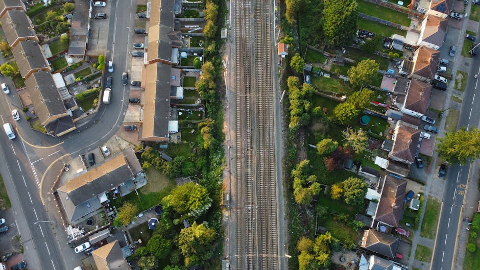 An aerial view of a residential neighbourhood showing a set of railway tracks running vertically through the centre, bordered by green trees and bushes on both sides. To the left, there are houses with pitched roofs, gardens, and parked cars along the narrow streets; some homes have driveways and fencing. To the right, more houses with gardens and trees are visible, with vehicles parked along the streets and a sidewalk. The scene is illuminated by natural daylight, capturing the process of home relocation or moving services that involve careful planning of furniture transport and parking management, as highlighted by Southwark Council rules for borough removals and parking permits, as referenced on manandvanborough.co.uk.
